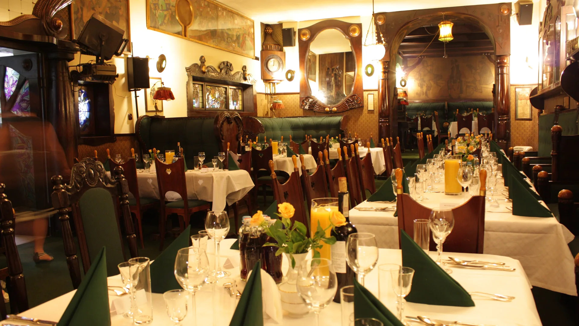 Long tables laid with white cloths and green napkins in a room of carved dark wood, mirrors and green benches at Jama Michalika.