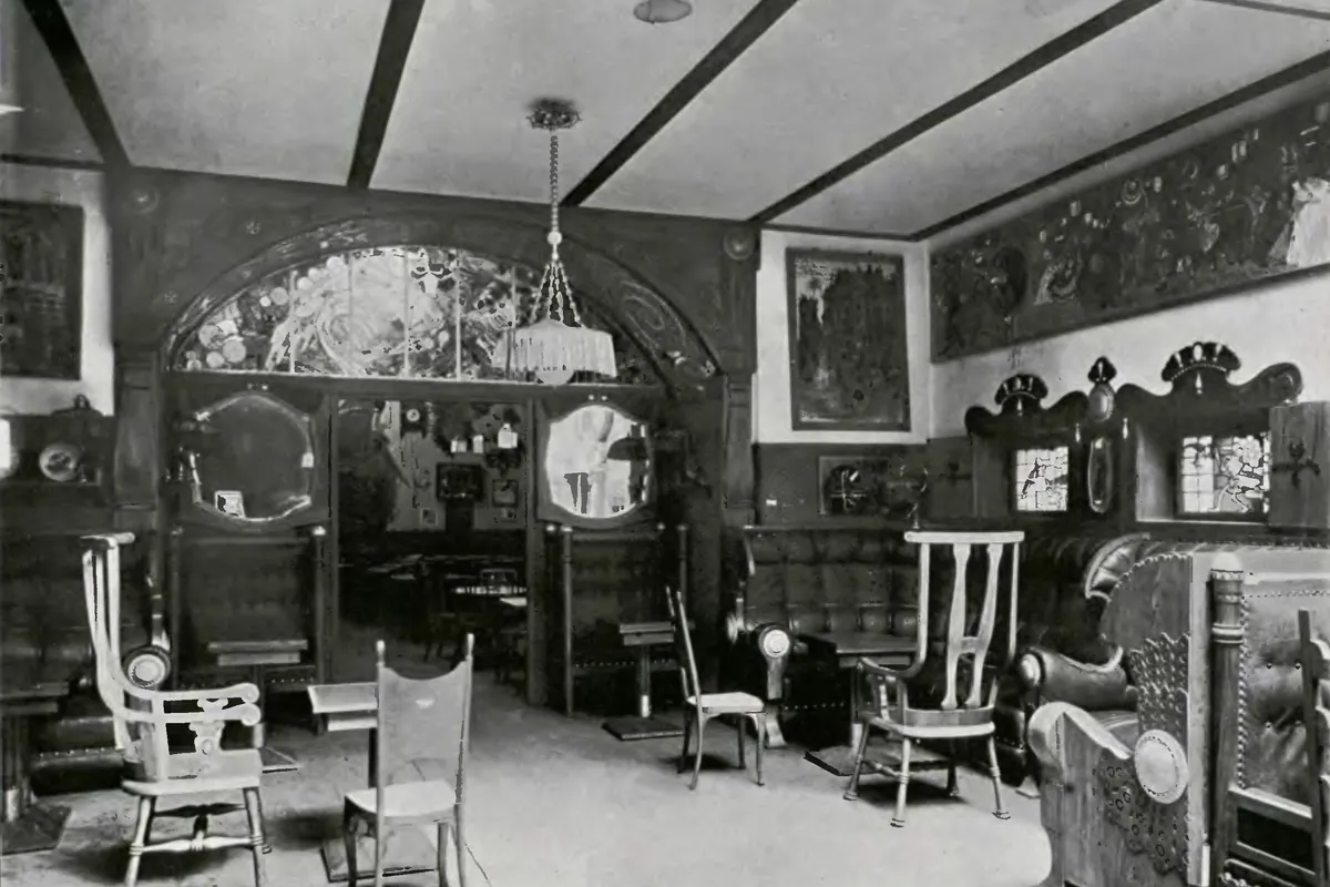 A black-and-white photograph of the café interior around 1911, with a stained-glass arch, round mirrors and light wooden chairs.